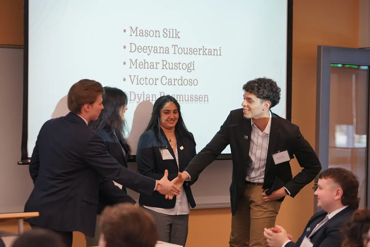 Two young men in business attire shake hands in front of two other people standing in front of a screen in a classroom.