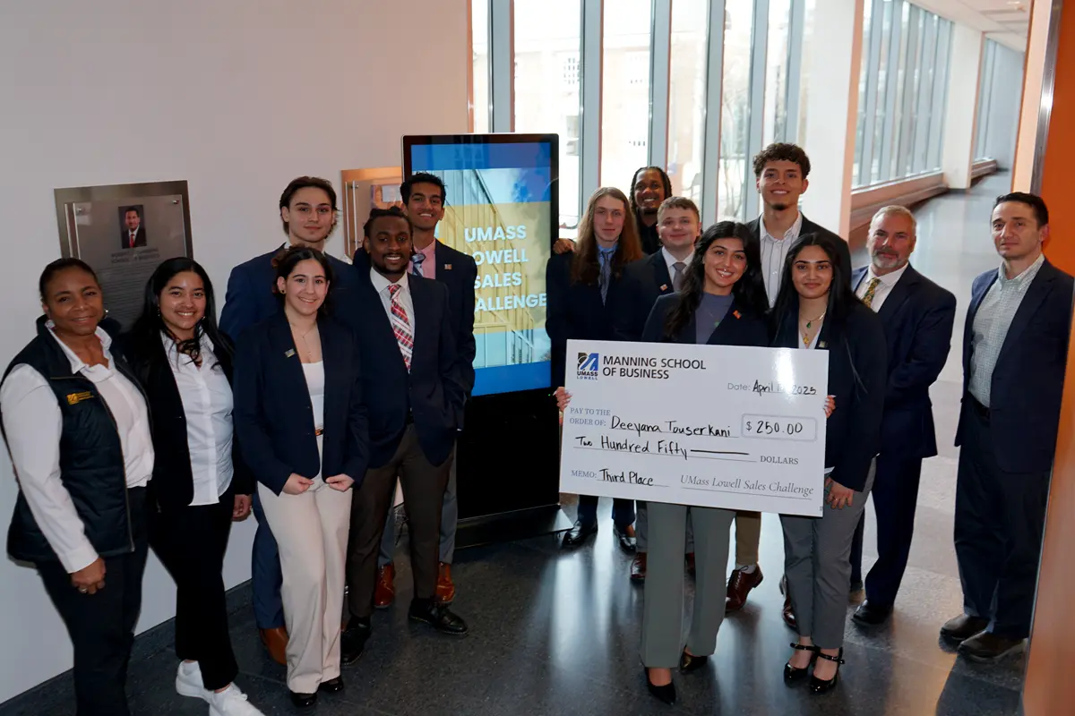 A group of 14 people pose for a photo in front of a monitor in a hallway. One of the people is holding a giant check. 