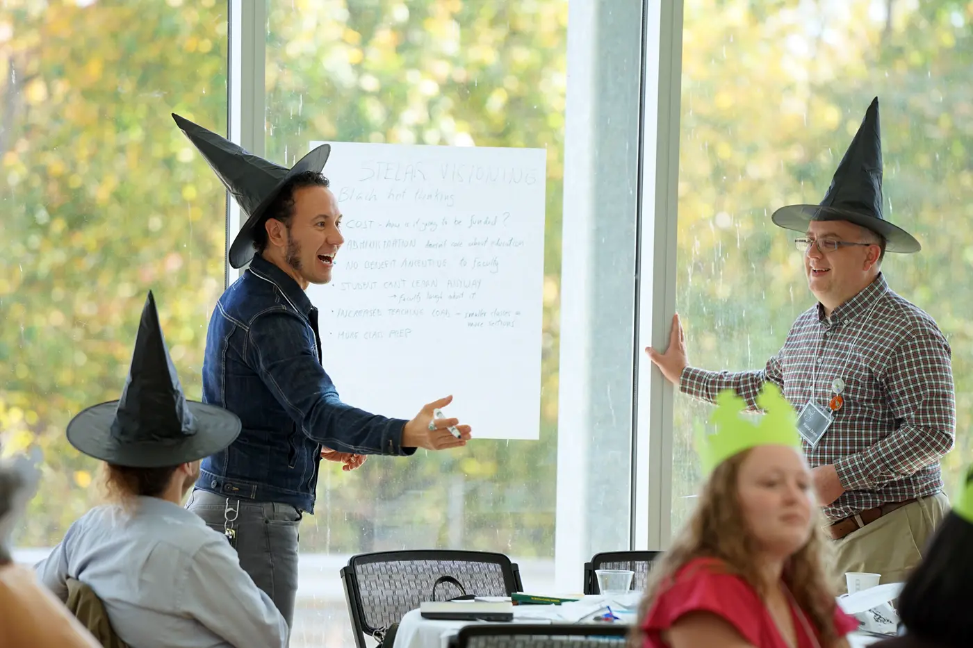 Two men wearing black witches hats stand next to a piece of paper taped to a window.