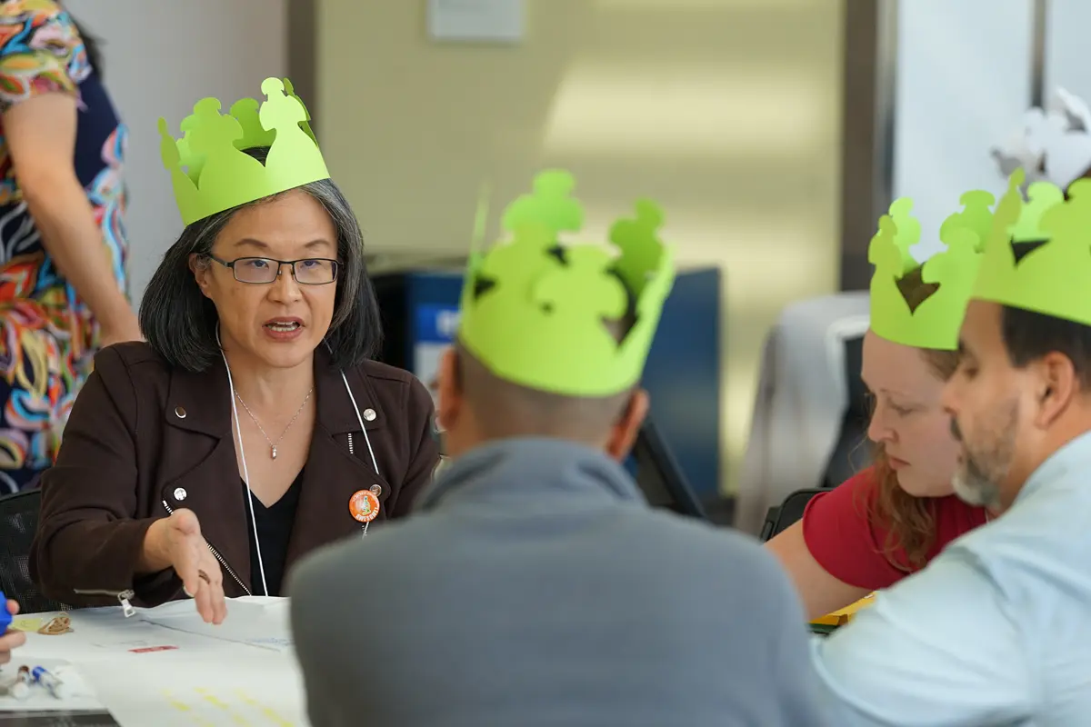 A woman wearing a bright green paper crown talks to three other people wearing the same crowns at a table.