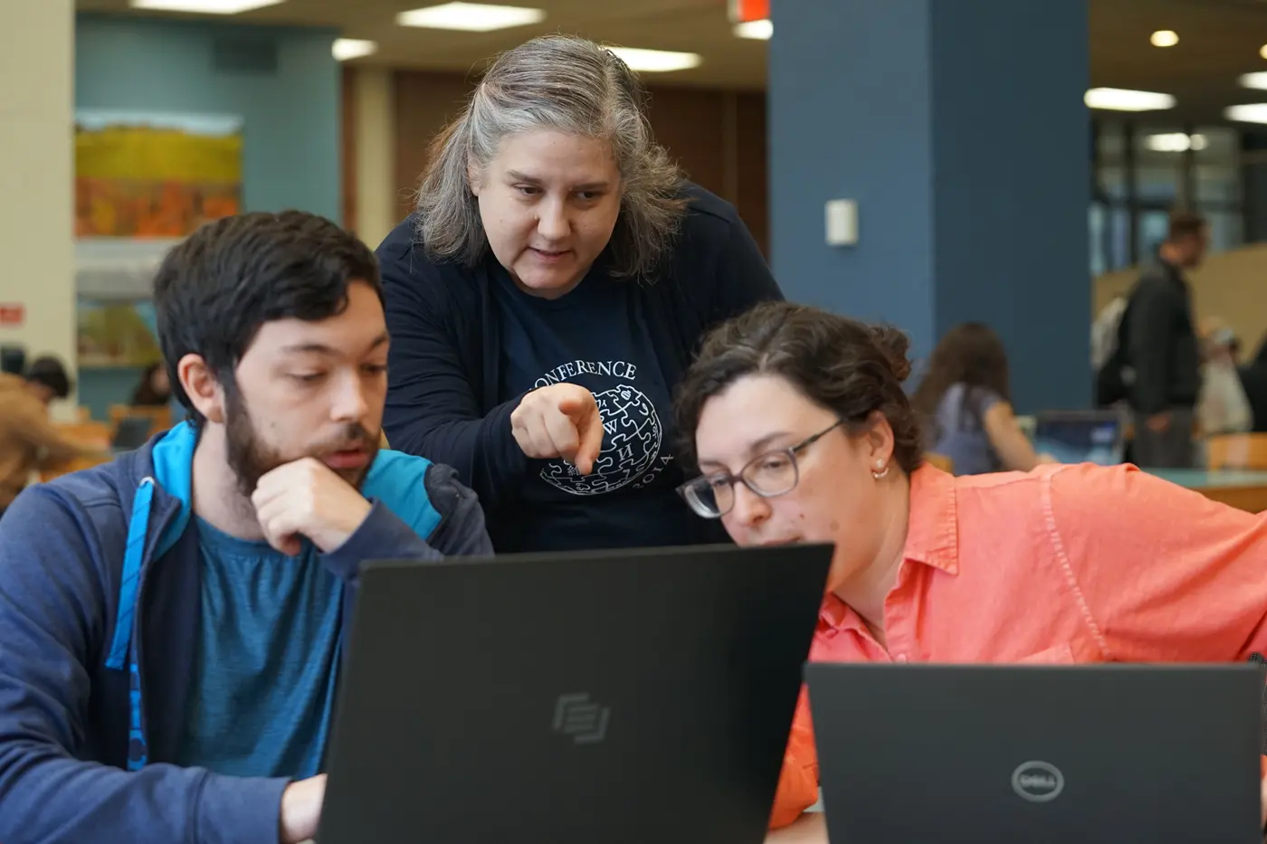 A standing person points at a laptop screen while two seated people look at the screen in a library.