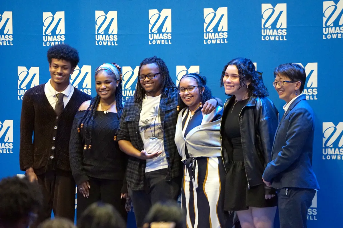 Five college students and a woman in a suit pose for a photo while standing in front of a blue backdrop on a stage.