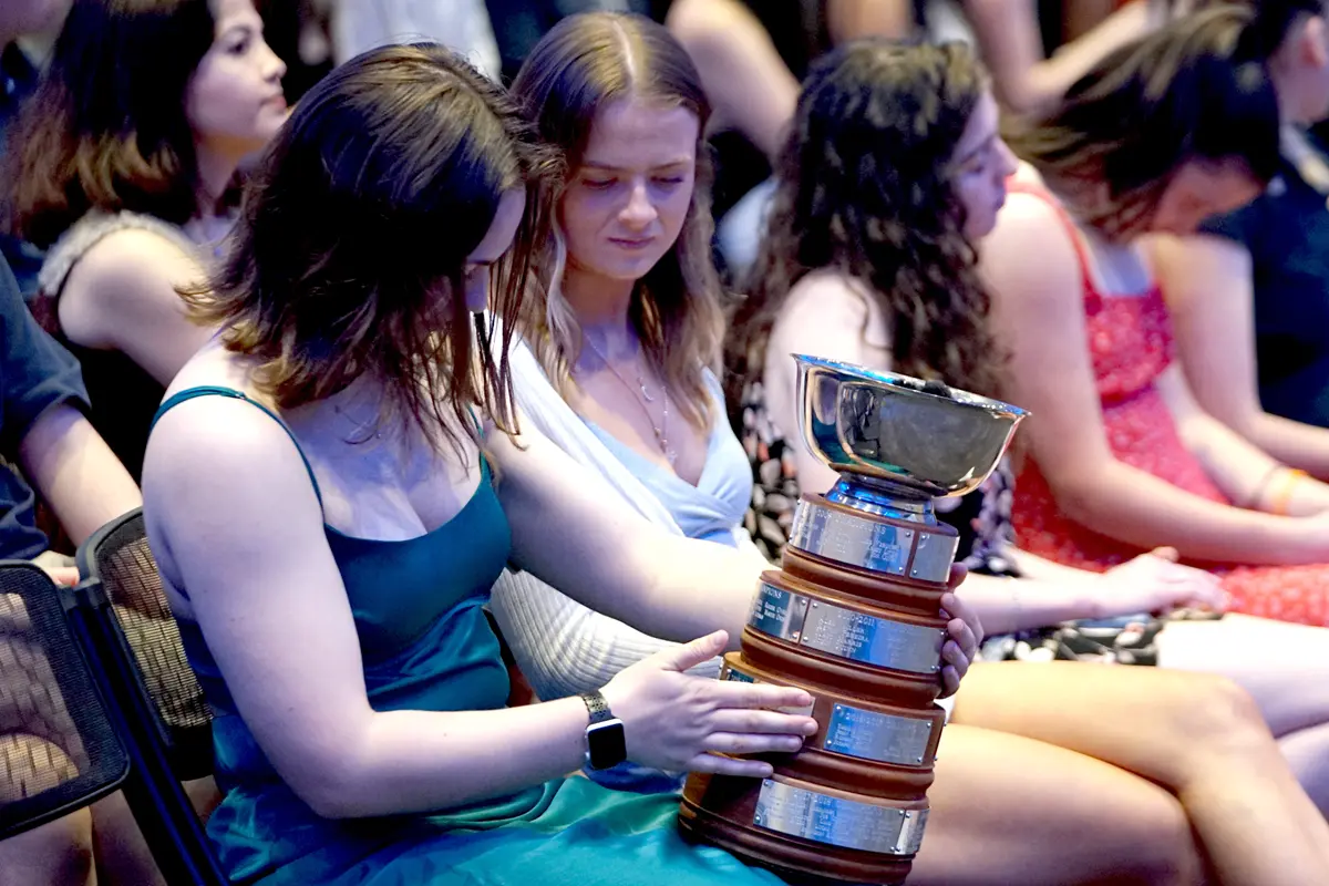 A young woman looks at a trophy she is holding on her lap while another woman looks on.