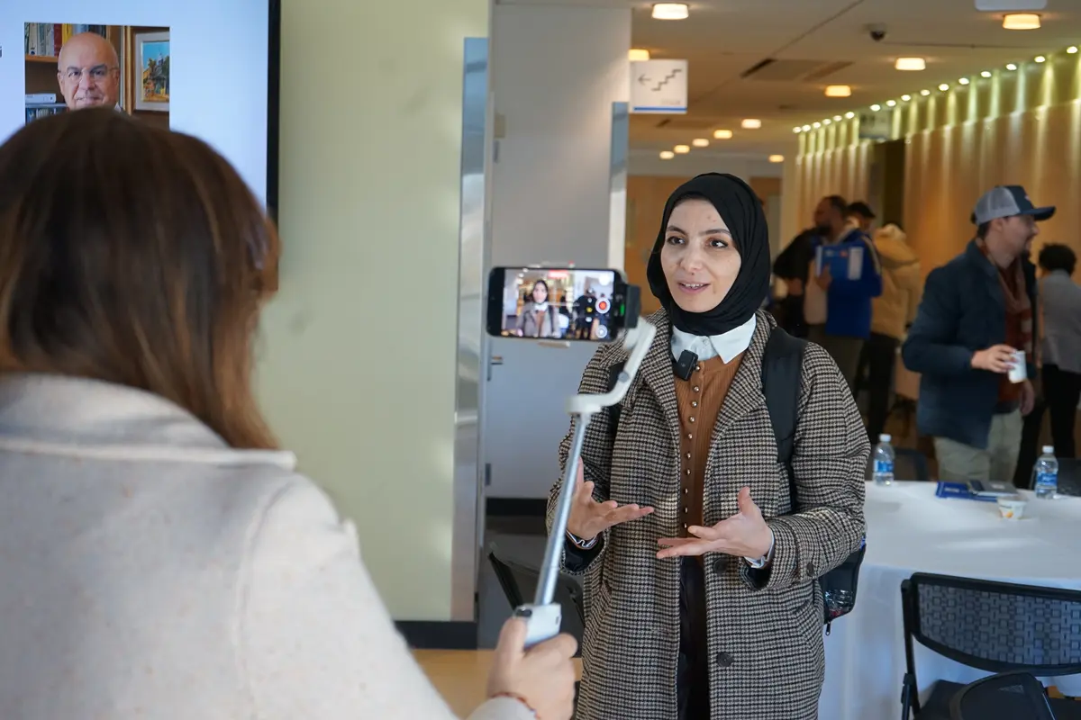 A woman is recorded while speaking by another woman holding a camera on a selfie stick.