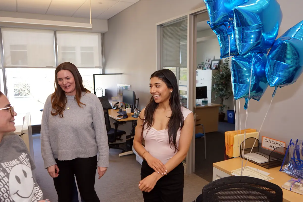 A young woman smiles while talking to people in an office with blue mylar balloons next to her.