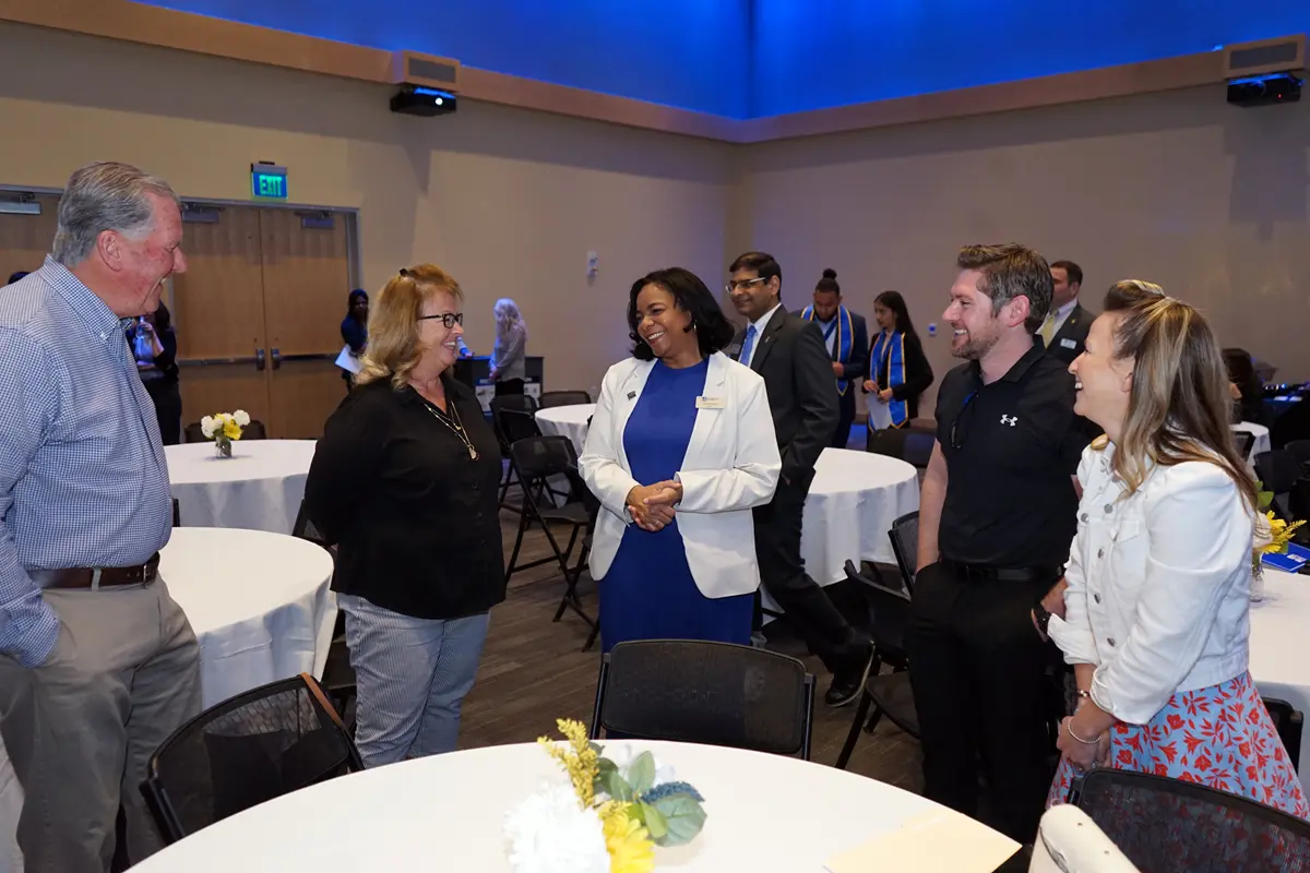 Two men and three women smile and chat while standing around a table in a ballroom.