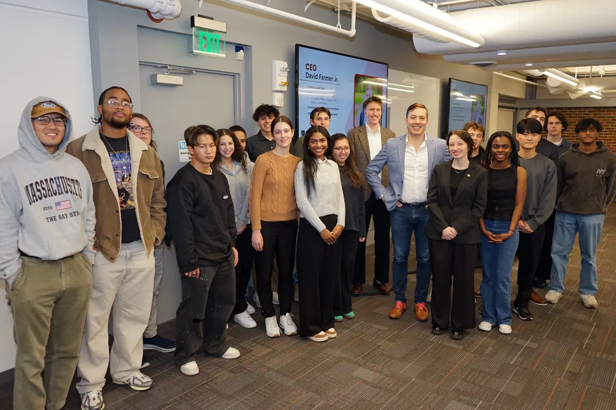 A group of college students pose for a photo with a man in a blue blazer in a room in front of a projection screen.