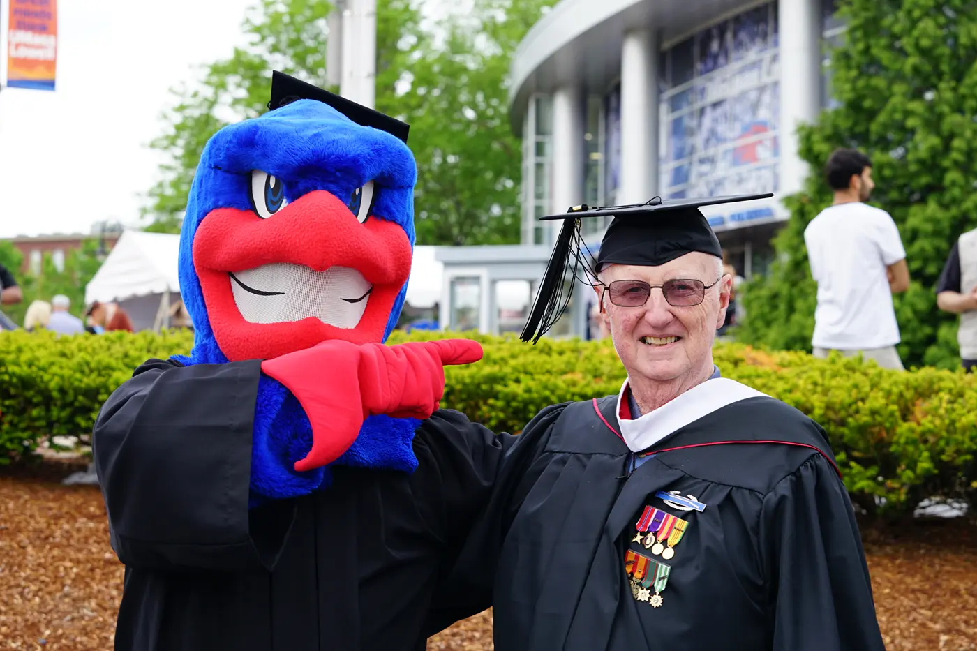 A blue bird college mascot points to a man in a graduation cap and gown. 