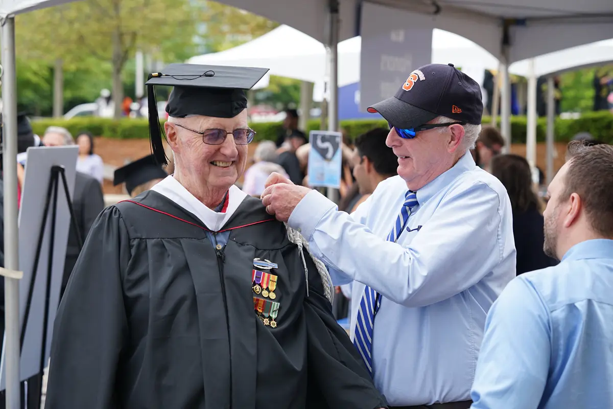 A man in a graduation cap and gown has a white cord attached to his shoulder by a man in a baseball cap and sunglasses.
