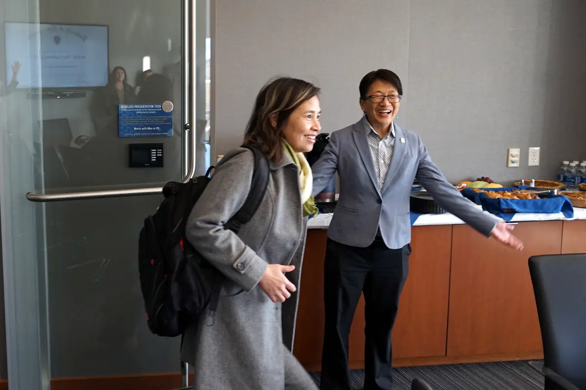 A woman enteres a room with a backpack over her shoulder while a woman in a blazer gestures with her arms behind her.