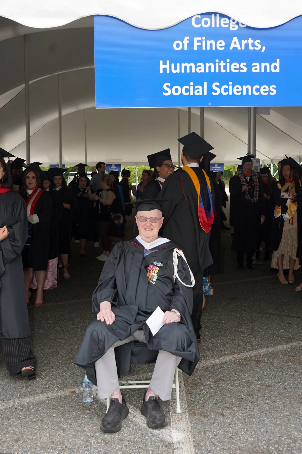 A man in a cap and town sits in a chair in front of a tent full of other college graduates.