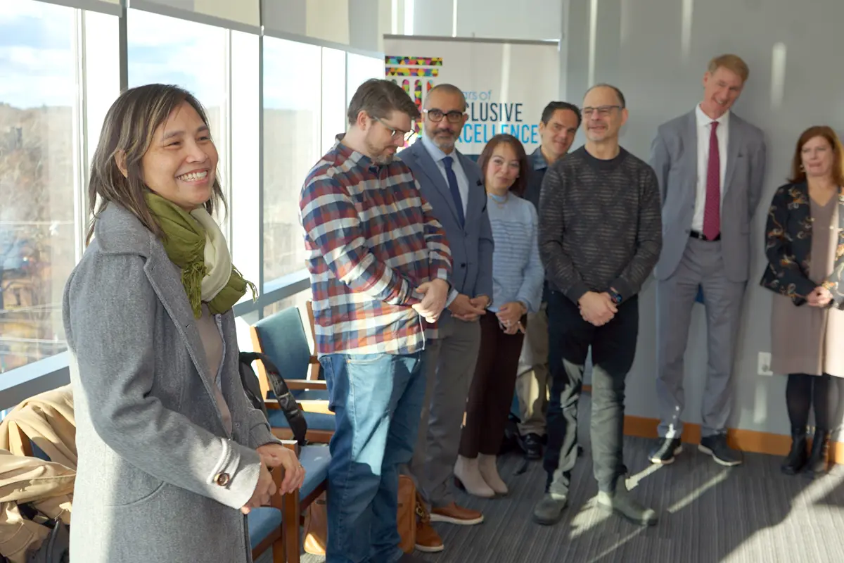 A woman smiles while standing in a room with a half dozen people facing her.