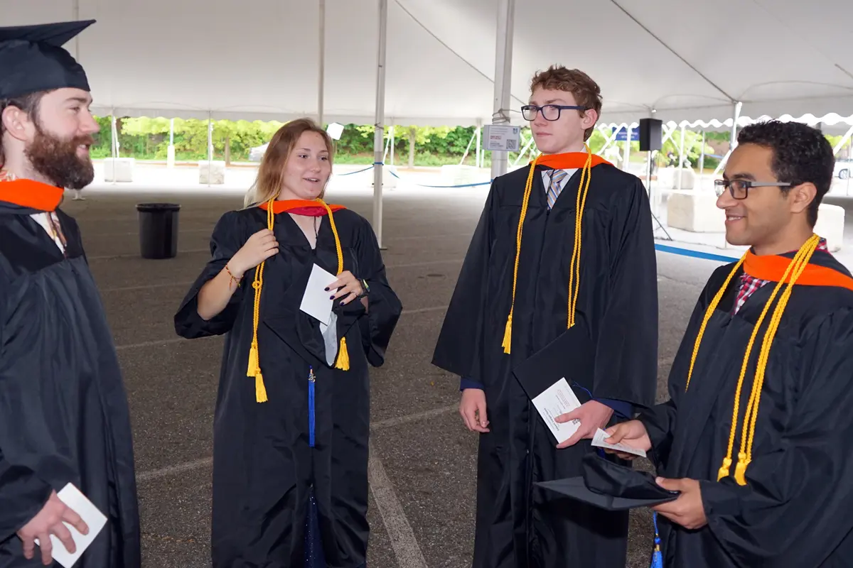 Four college graduates in caps and towns stand under a white tent talking to each other.