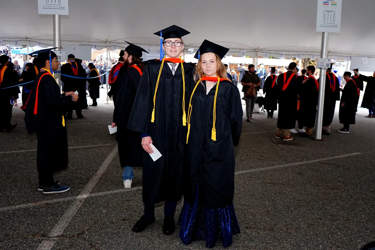 Two college graduates in caps and gowns pose for a photo under a tent.