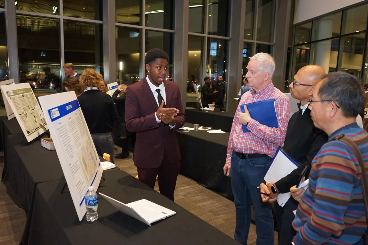 A young man in a suit presents a poster to three men in a building lobby.