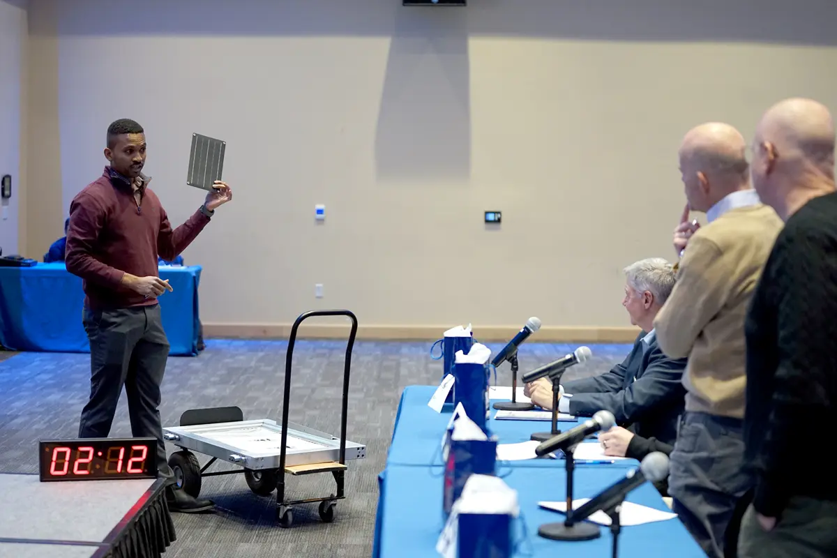 A man holds a small solar panel while facing a table of judges.