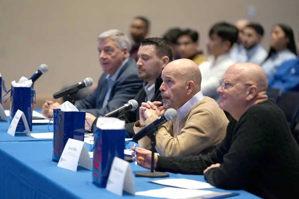 A man seated at a long table with three other men asks a question into a microphone.