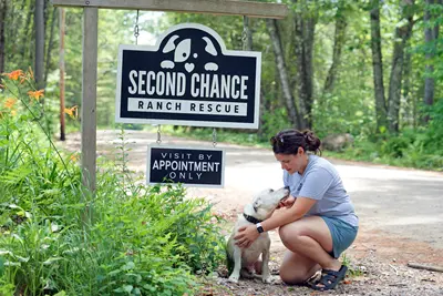 A woman in shorts and a T-shirt pets a white dog near a sign that says Second Chance Ranch Rescue Visit by Appointment Only on the side of a road.