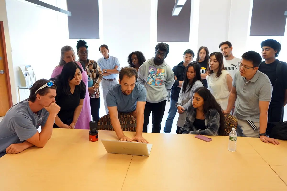 A group of students gather around a table as a young man shows them something on his laptop.