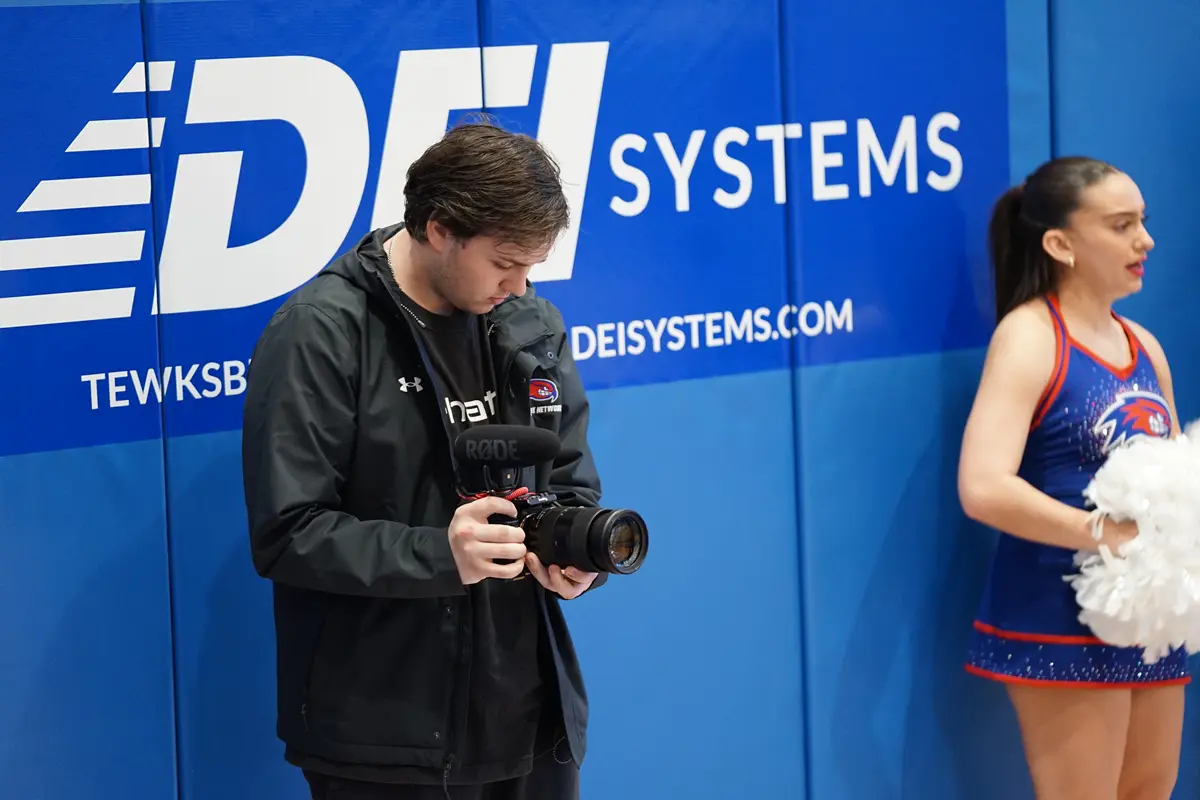 A young man in dark clothes holds a digital camera while standing next to a cheerleader on the baseline of a basketball court.