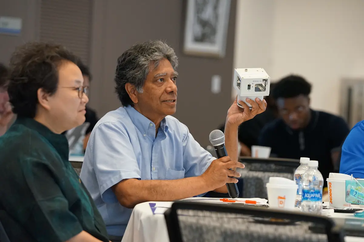 A man in a blue shirt holds up a small satellite while sitting at a table.