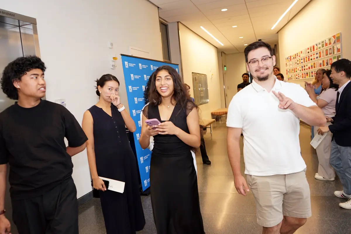 A man in a white shirt gestures a peace sign while three other people look on in a hallway.