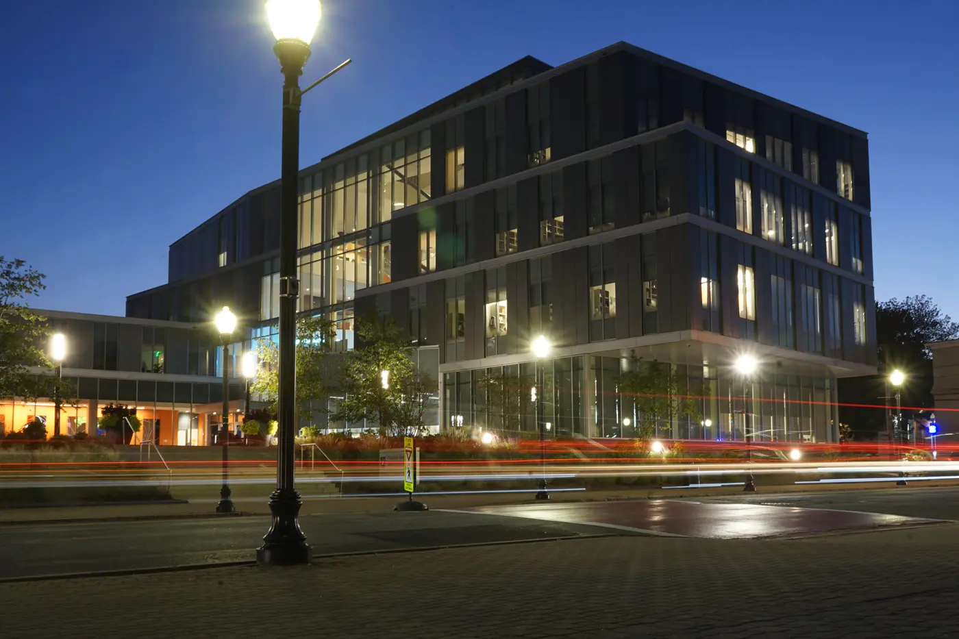 Car headlights blur past a business school building at dusk.