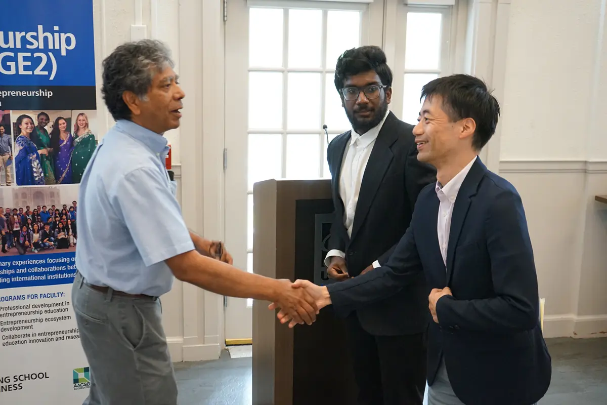 A man in a light blue shirt shakes hands with a man in a suit while another man in a suit looks on.