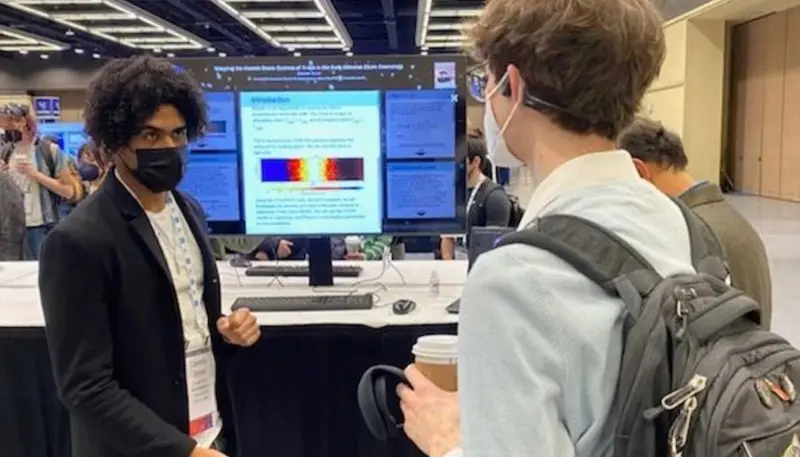 UMass Lowell physics student Dashon Jones talks with a student while standing before a computer screen displaying data.