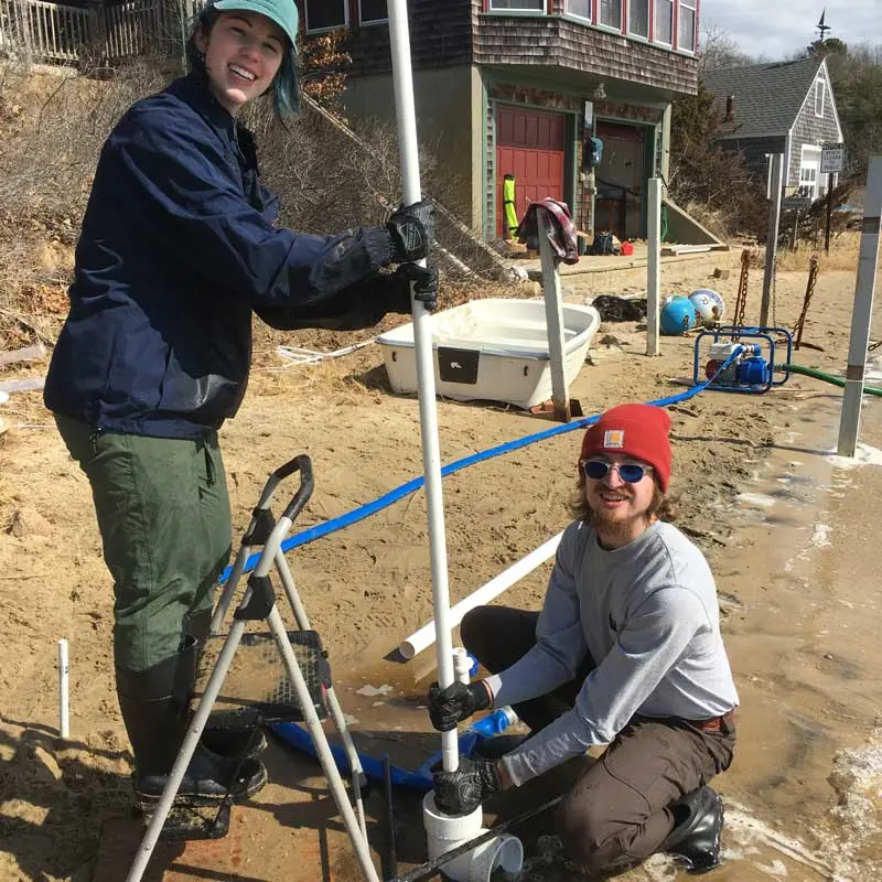 Two students use equipment to conduct tests along the ocean shoreline.