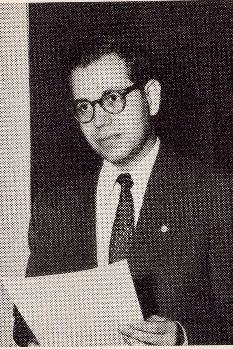 A man wearing glasses and a suit holds a piece of paper and looks off to the left.