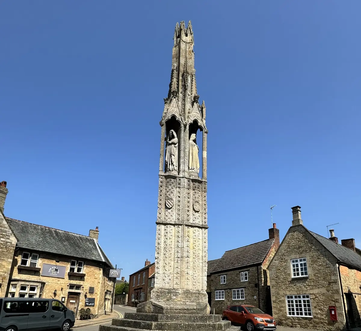 Medieval Stone Eleanor Cross in town center surrounded by buildings and cars.