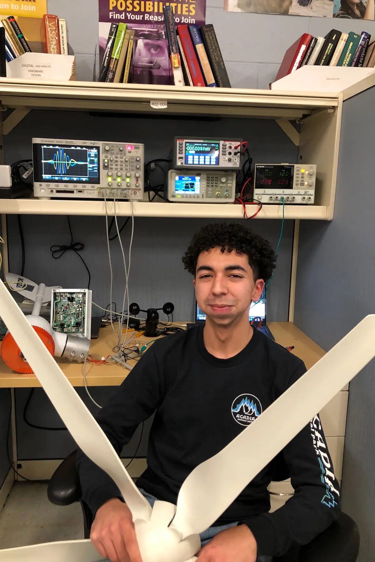 Driss Bourzgui sitting and holding a fan propeller in front of his desk stacked with equipment.