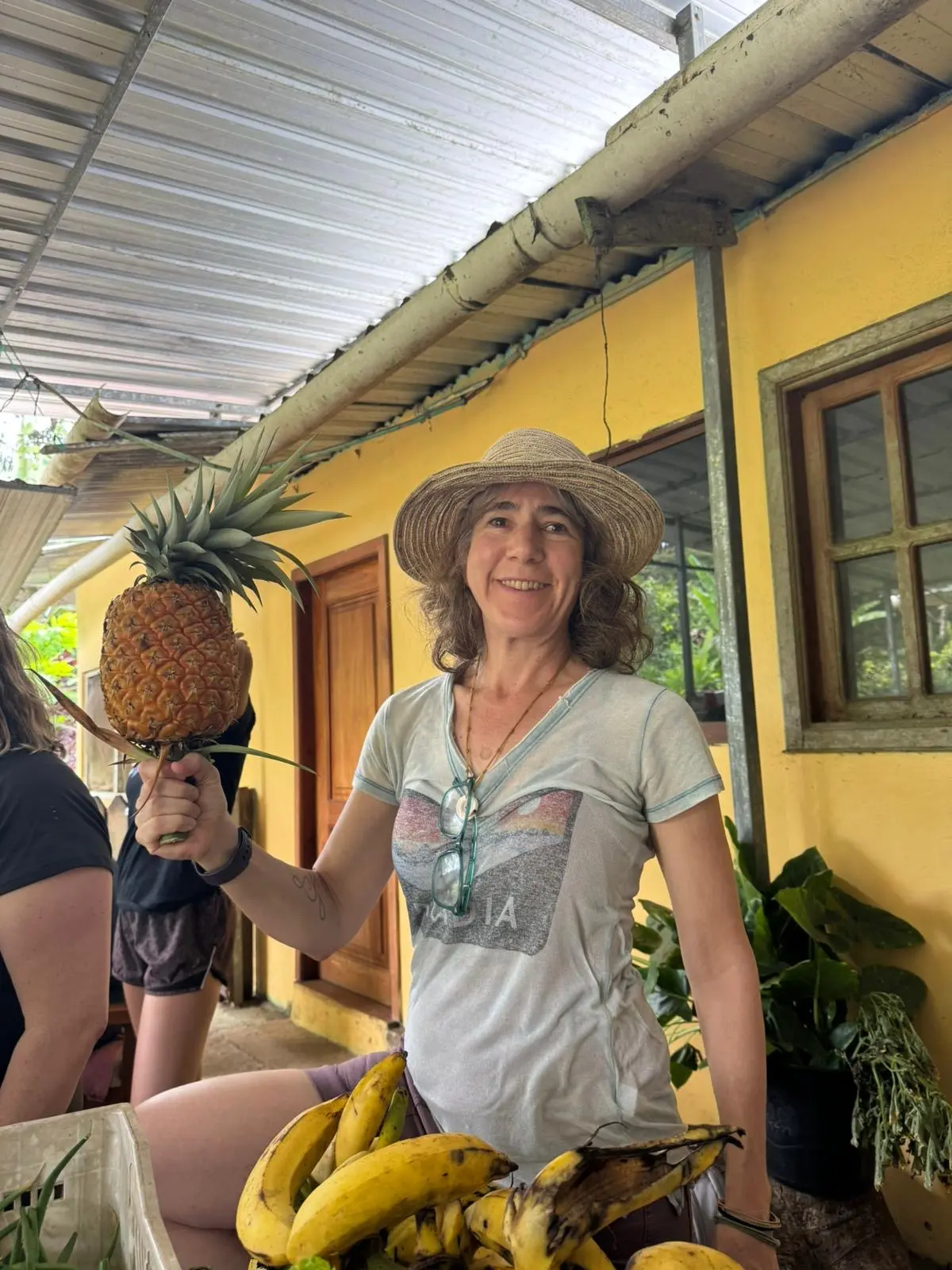 A woman in a T-shirt and a hat poses for a photo while holding a pineapple.