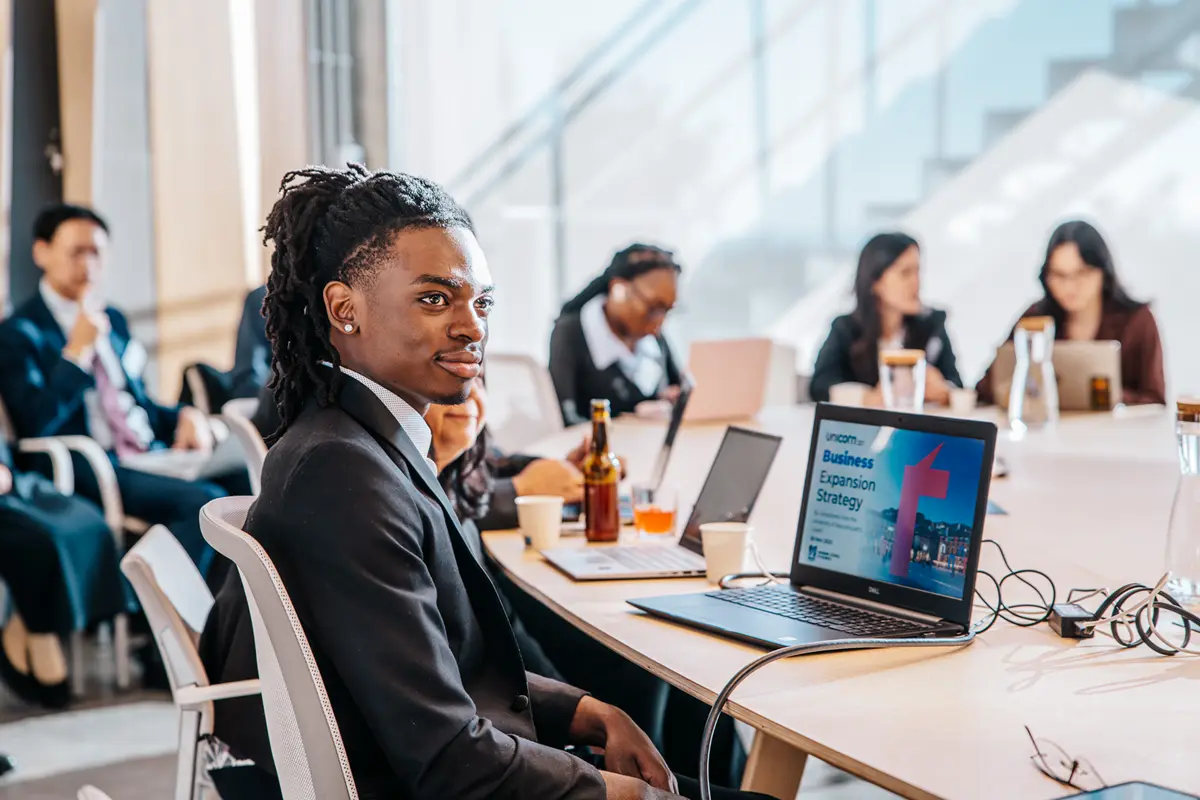 A young man in a suit sits at a conference table with a laptop.