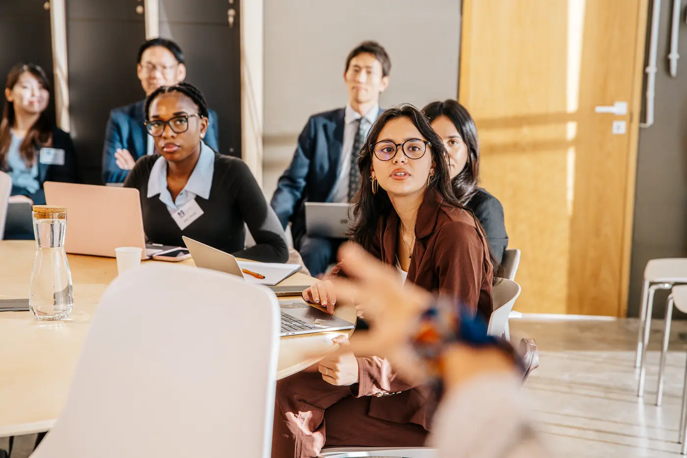 A half dozen men and women in business attire listen to a speaker while seated in a conference room.