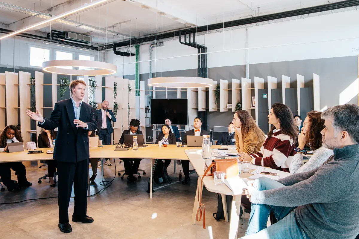 A young man in a suit makes a presentation while standing in front of a dozen people seated at tables in a room.