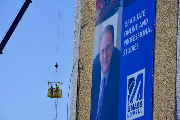 Workers suspended in a basket from a crane behind a GPS sign