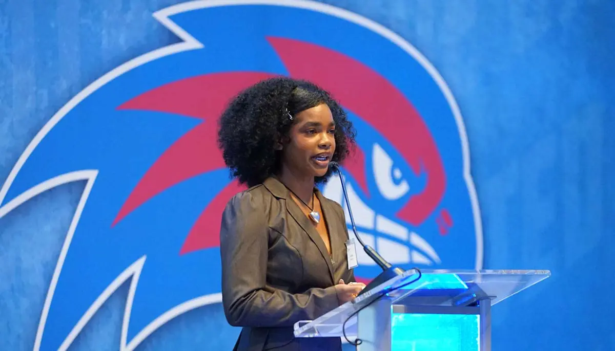 Gabriella Monteiro speaks at a lectern with the UMass Lowell logo in the background.