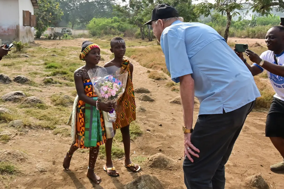 Two youngsters present a bouquet of flowers to a man outside while a man takes a photo.