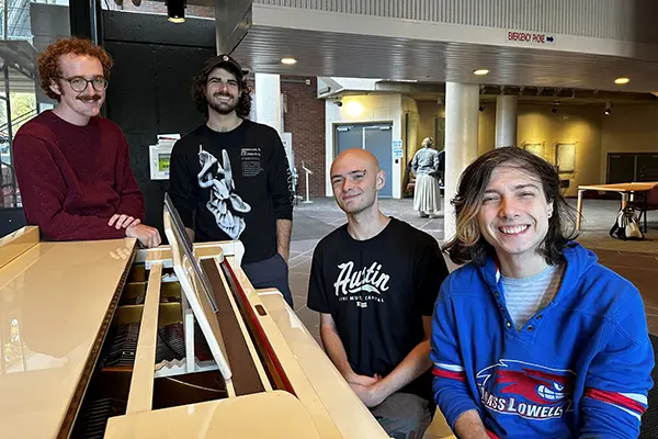 The Ghouls band members sit and stand around a piano in Moloney Hall