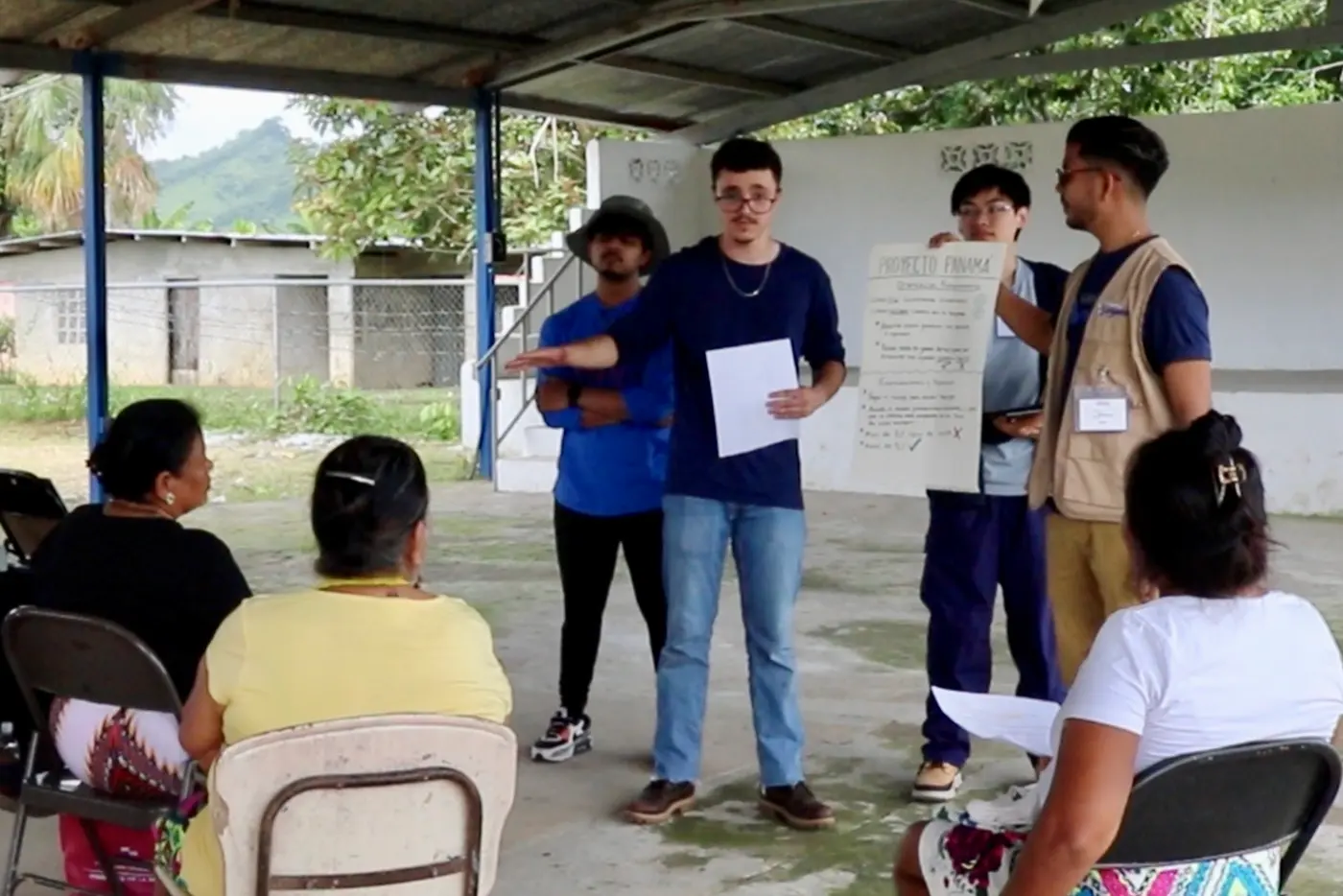 A young person makes a presentation to several seated people while a person holds a piece of paper and two others look on.