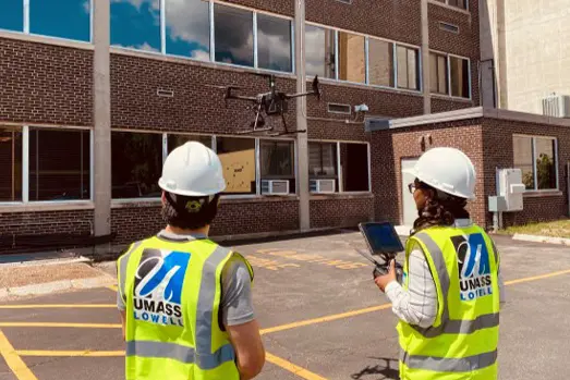 Two researchers wearing UMass Lowell vests watch a drone fly.