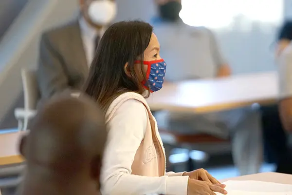 A woman with dark hair wearing a face covering sits in a classroom