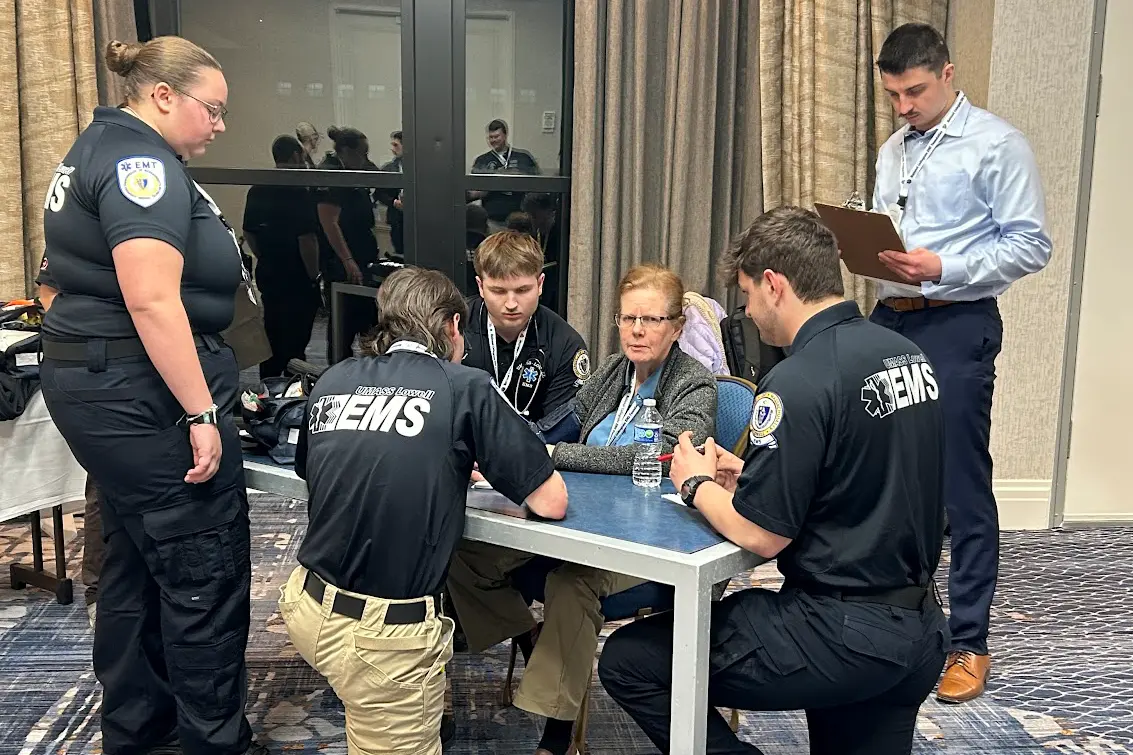 Four emergency medical workers talk to a woman who is playing the role of a patient while a person with a clipboard looks on.
