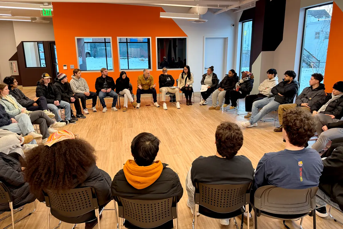 A group of about 20 people sit in a circle of chairs in a room with a wood floor.