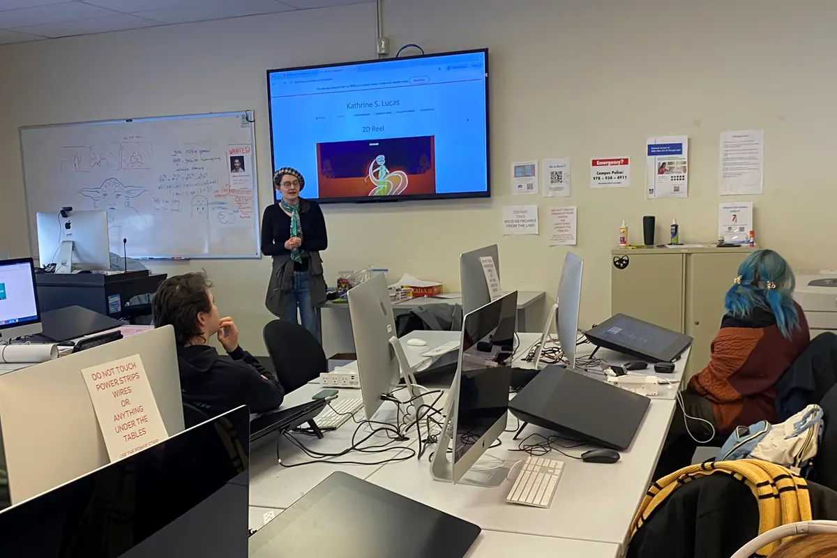 A young person stands next to a monitor while speaking to a college art class.