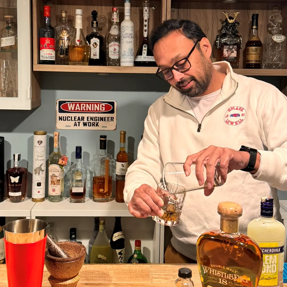 A man with dark hair and glasses mixes a cocktail while standing behind a bar.