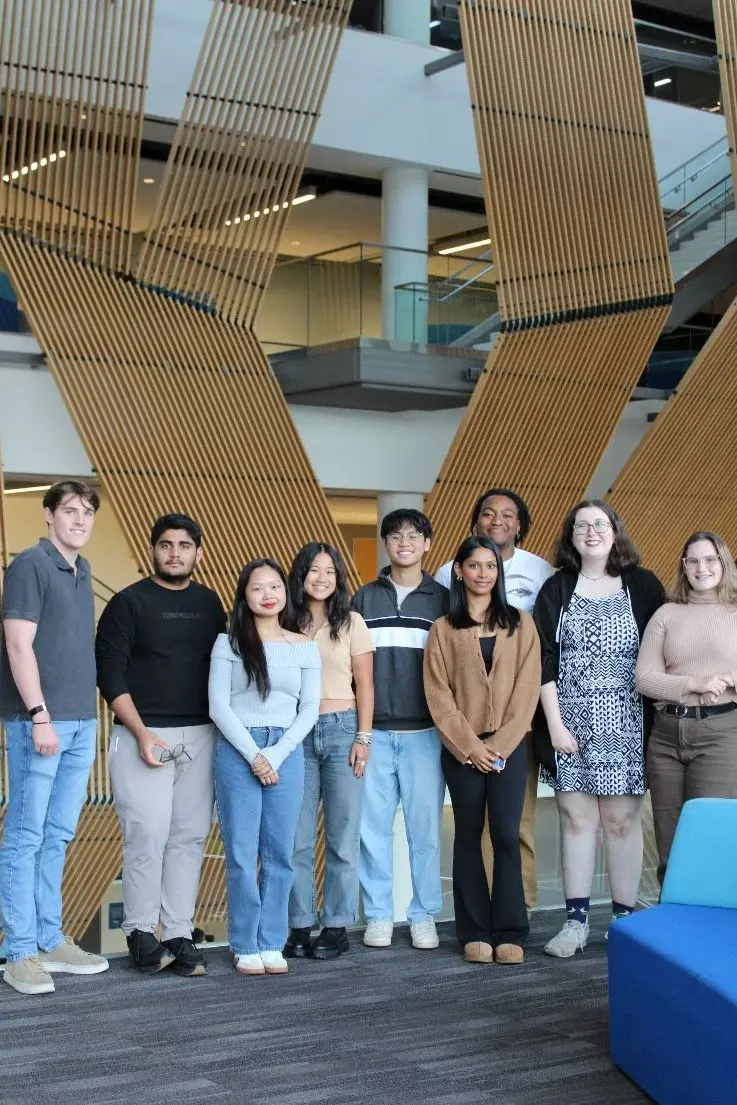 Nine students stand smiling at camera with a wooden slat structure behind them