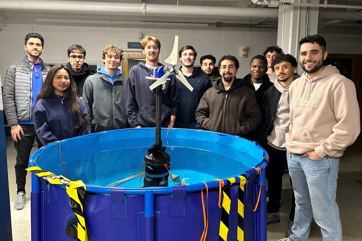 A dozen college students pose for a photo standing around a small blue pool with a prototype wind turbine floating in it.